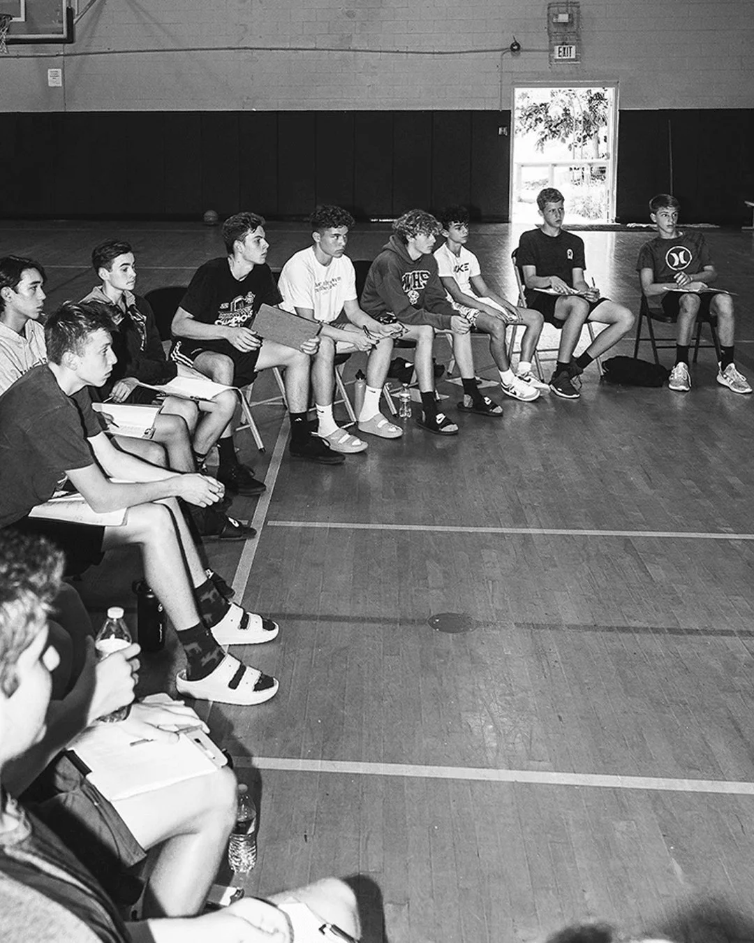 A group of young boys sitting on chairs in a gymnasium, appearing to be in a meeting or discussion, some holding notebooks or papers, with a basketball court visible in the background.