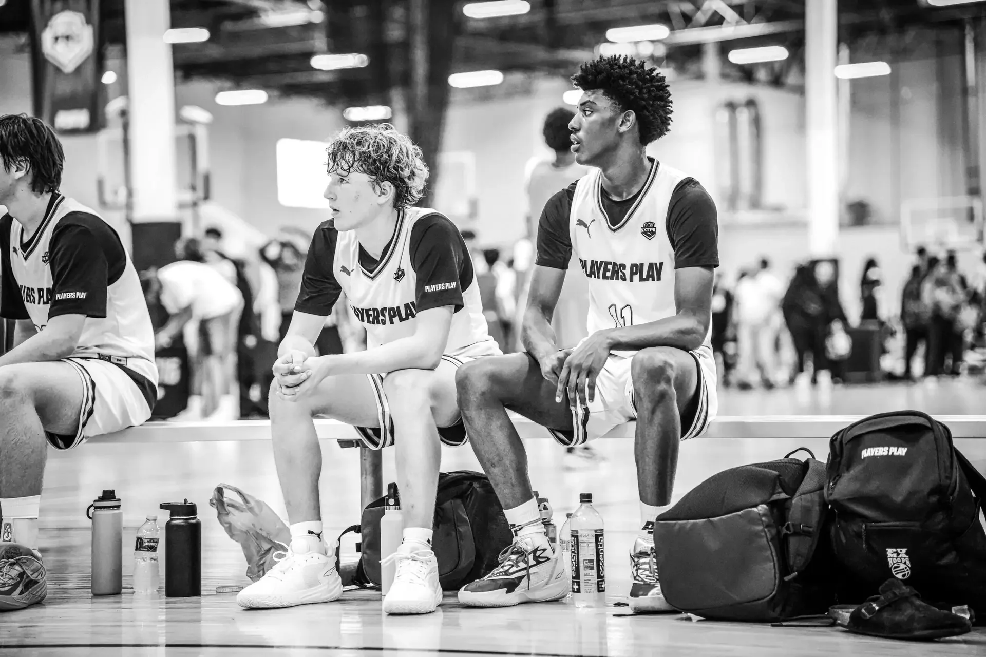 Two male basketball players sitting on a bench during a game, wearing jerseys with the text 'PLAYERS PLAY'.