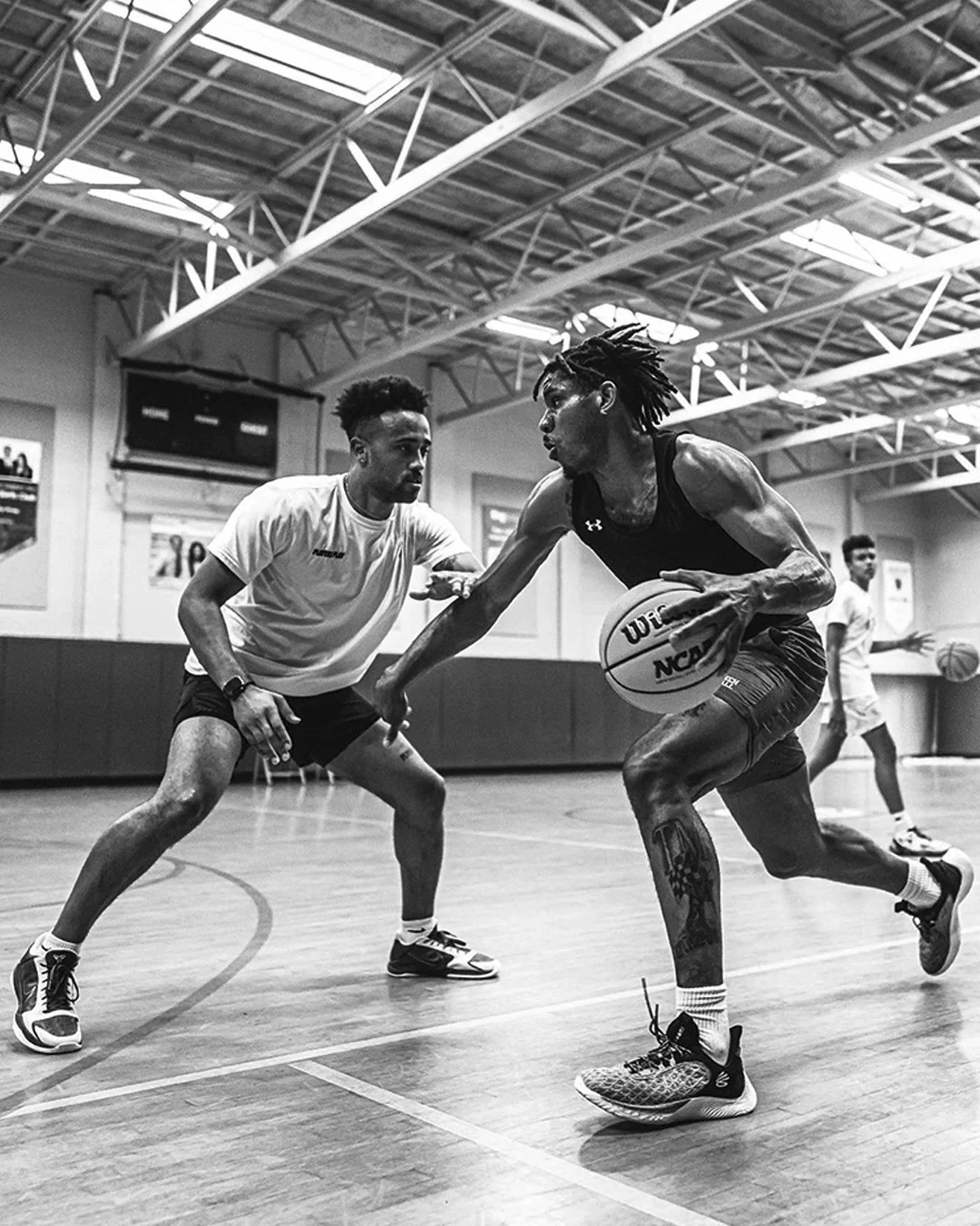 Two basketball players are competing on an indoor court, one holding a basketball and dribbling while the other tries to defend. There are two additional players in the background and the gym has a high ceiling with exposed beams.