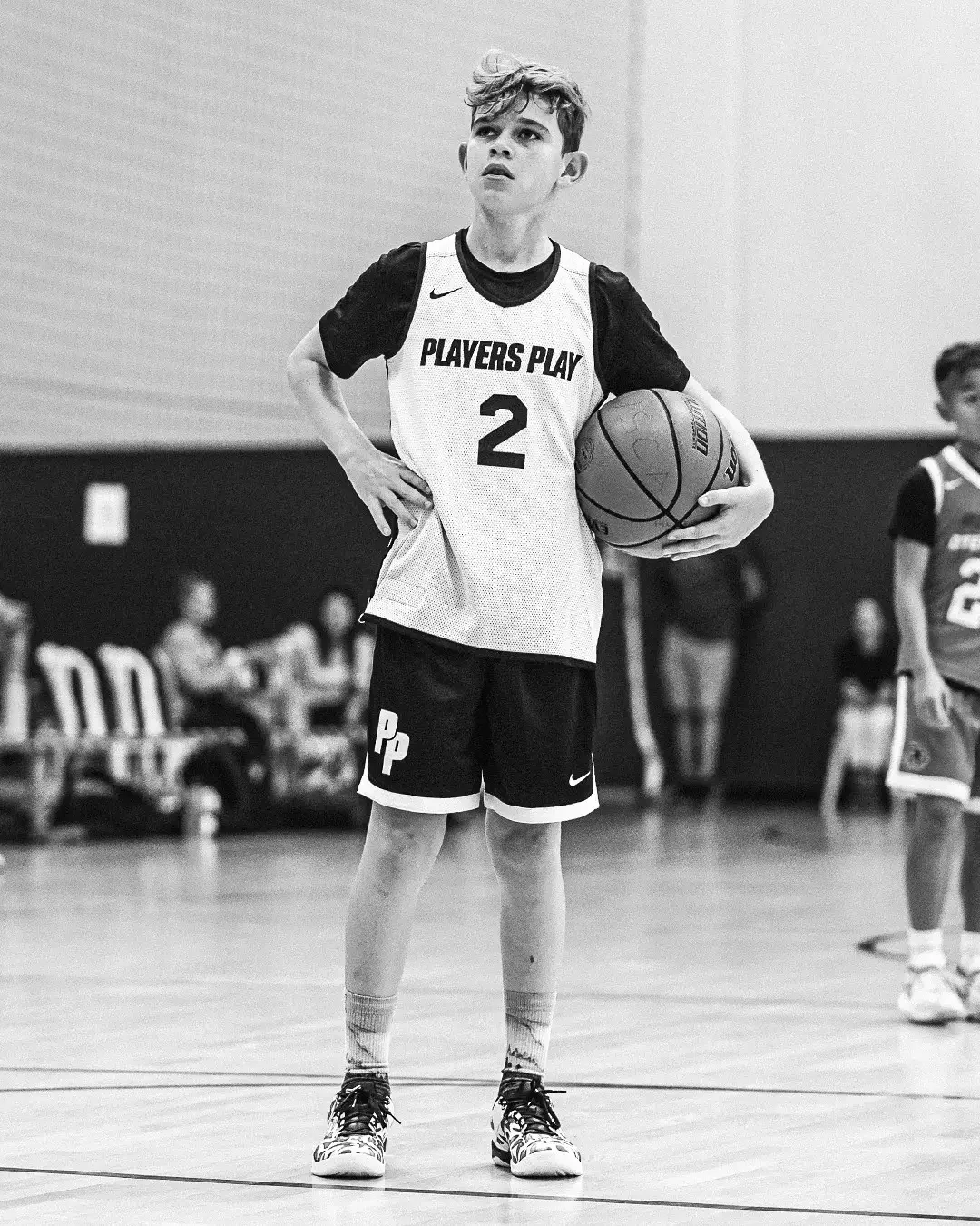 A young male basketball player holding a basketball on the court, wearing a jersey with 'PLAYERS PLAY' and the number 2, in a game setting.