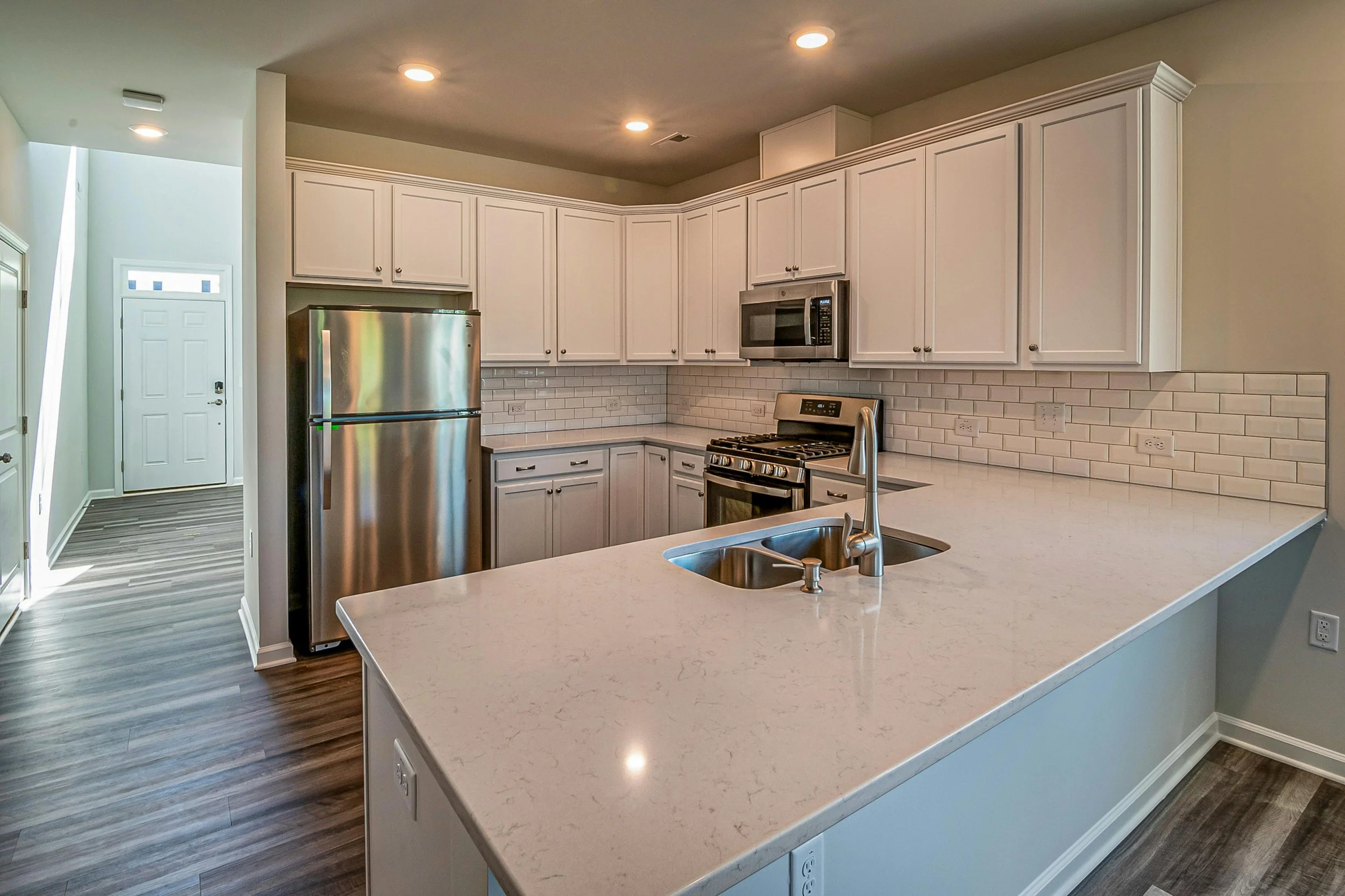 Modern kitchen with white cabinets, stainless steel refrigerator, microwave, gas stove, and a large white countertop island with a sink, hardwood flooring, and recessed lighting.
