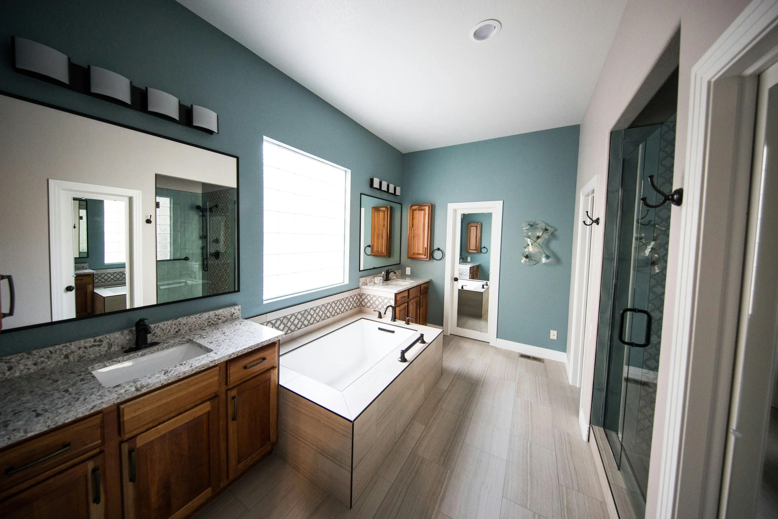 Modern bathroom with a large mirror, granite countertop, wooden cabinets, bathtub, glass shower, and decorative wall art.