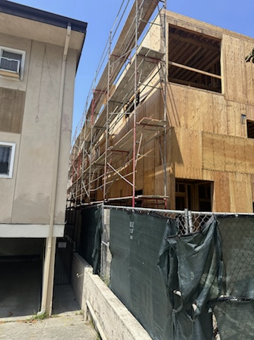 Construction site with scaffolding and unfinished wooden building next to a concrete building, with a chain-link fence in the foreground.