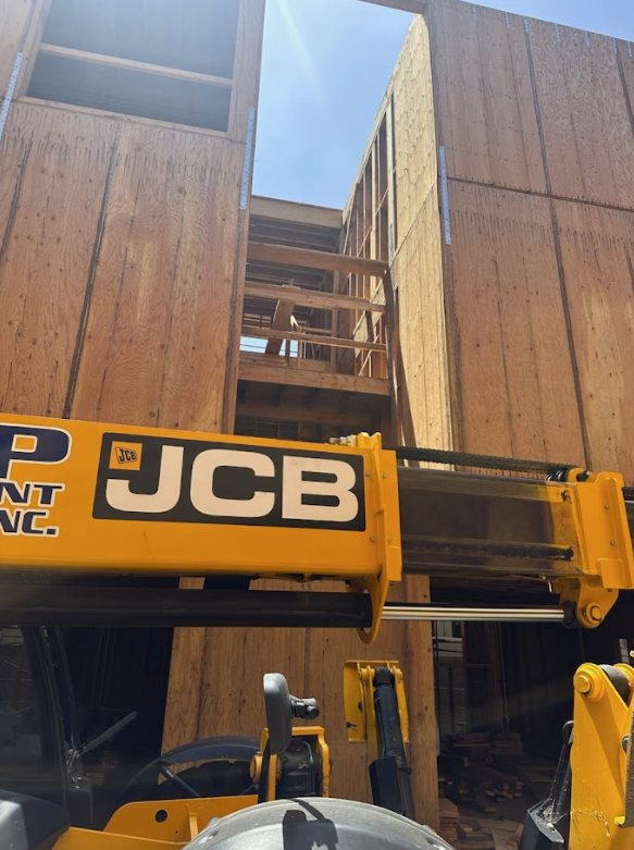 Construction site with wooden framing and a JCB crane lifting materials. Part of a building under construction with clear blue sky.