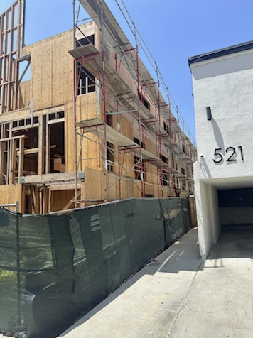 Construction site with a multi-story building under construction, scaffolding, and wooden framework next to a white building with the number 521 and an attached garage.