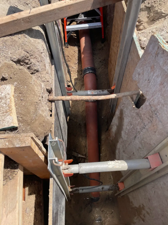 Construction site showing a large brown pipe installed in an excavation, with metal supports and framing around it.