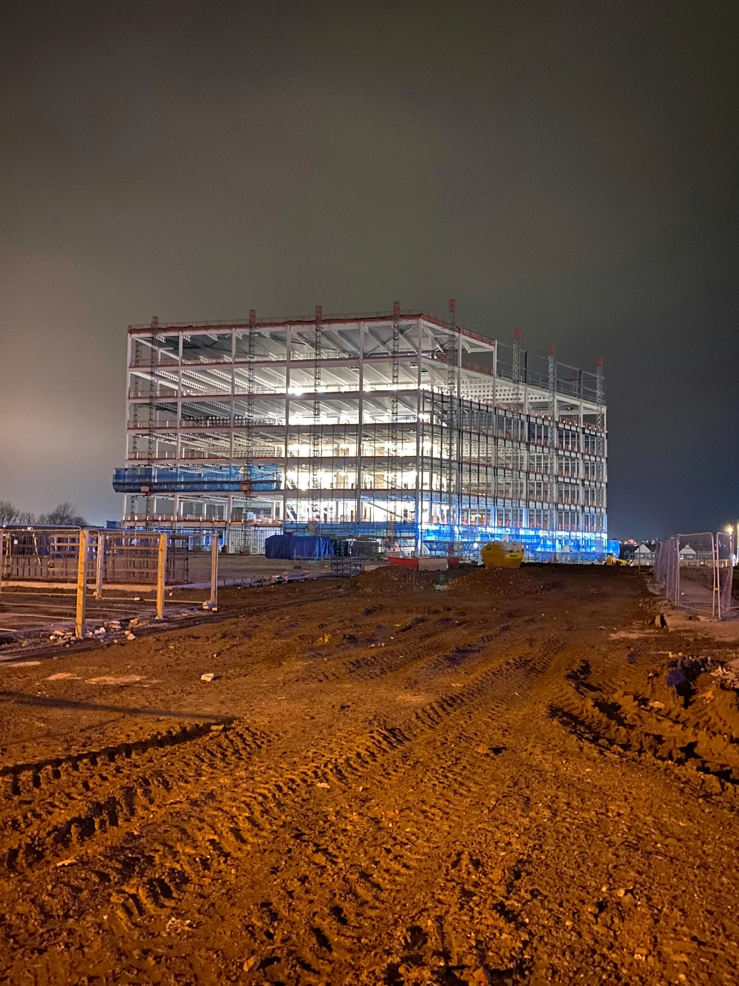 Construction site at night with a multi-story building under construction, scaffolding around the structure, illuminated from inside, with dirt ground and construction equipment in the foreground.