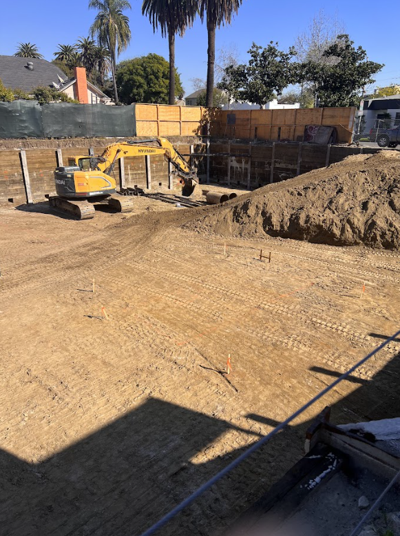 Construction site with a small yellow Hyundai excavator and a pile of dirt, surrounded by wooden fencing and trees in the background.