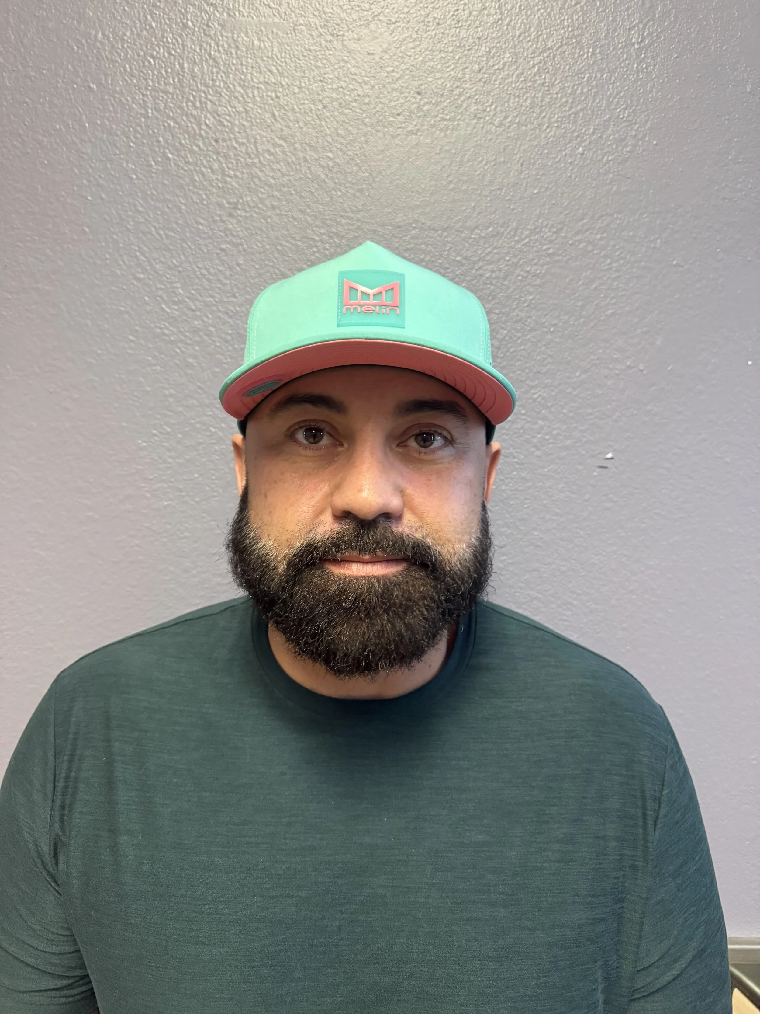 A man with a beard wearing a green T-shirt and a teal and pink baseball cap against a plain gray wall.