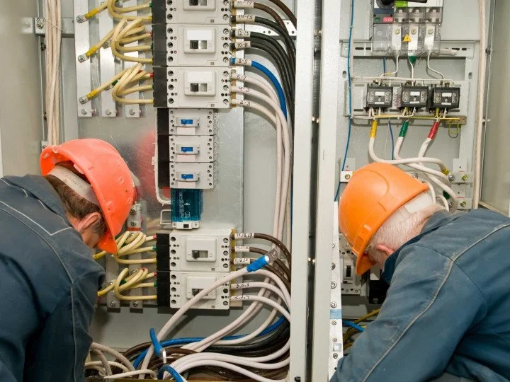 Two electricians wearing orange hard hats working inside an electrical control panel with wires and circuit breakers.