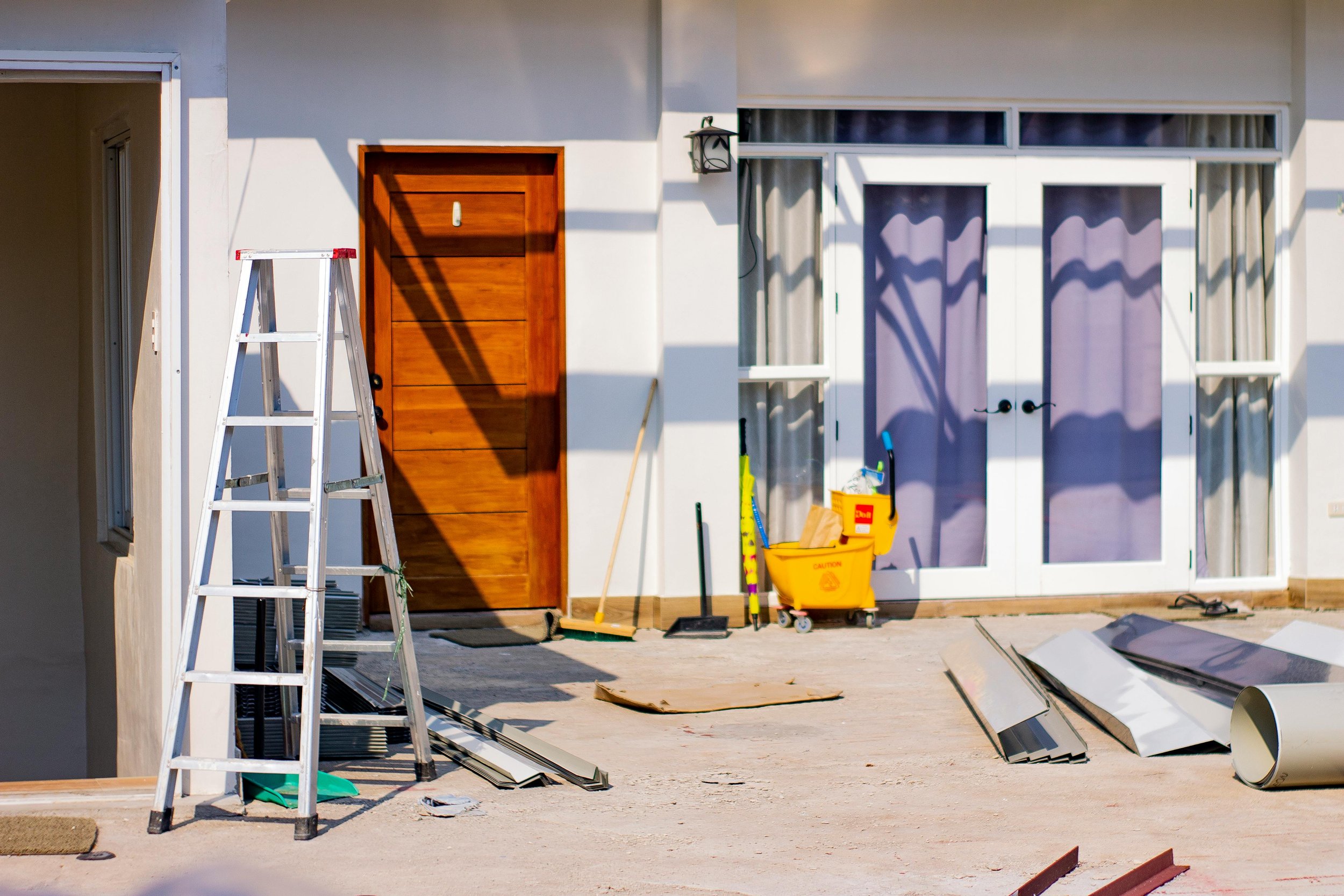 Exterior of a house under construction, showing a ladder, cleaning tools, and building materials in front of the door and window.