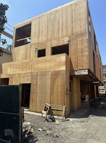 Construction site of a wooden building with unfinished exterior walls, open window and door spaces, and construction materials and scaffolding nearby.