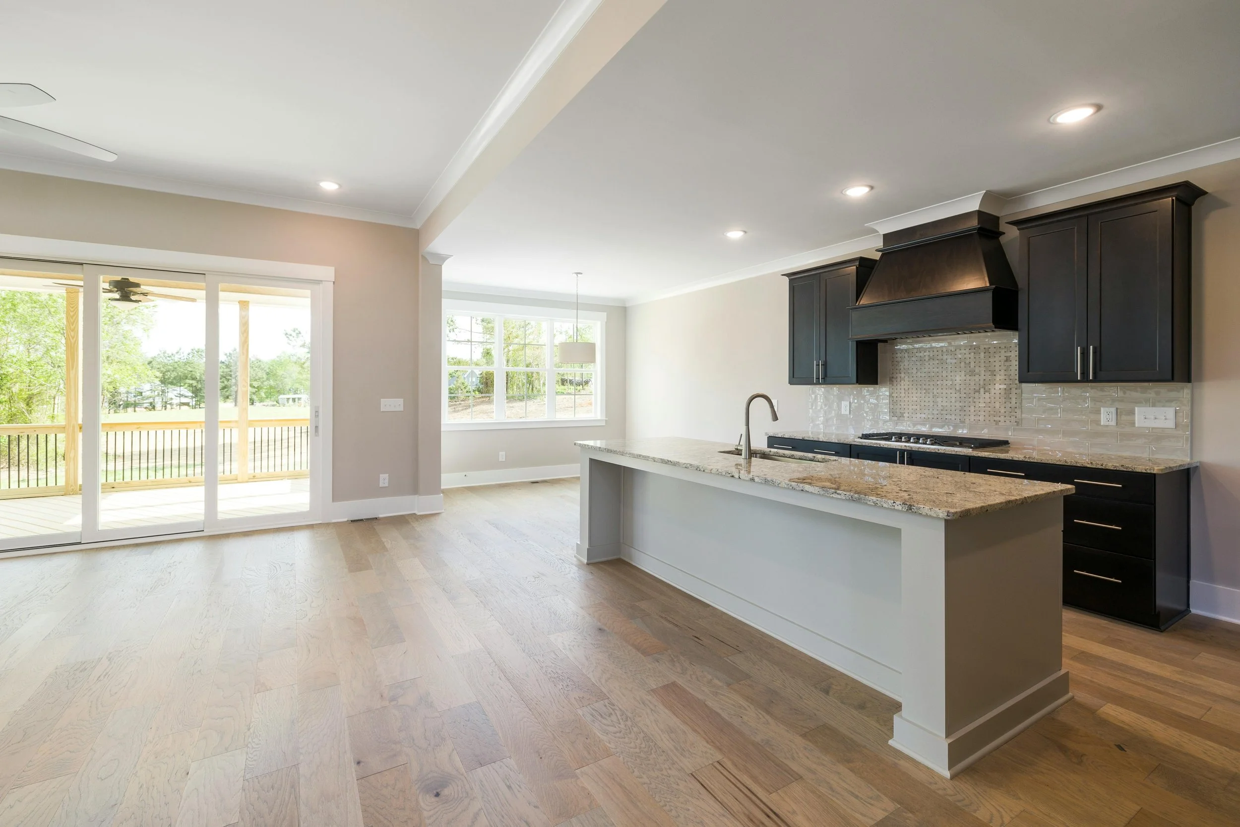 Open-concept kitchen with black cabinets, granite countertops, and a glass sliding door leading to a deck, with hardwood floors and large windows providing natural light.