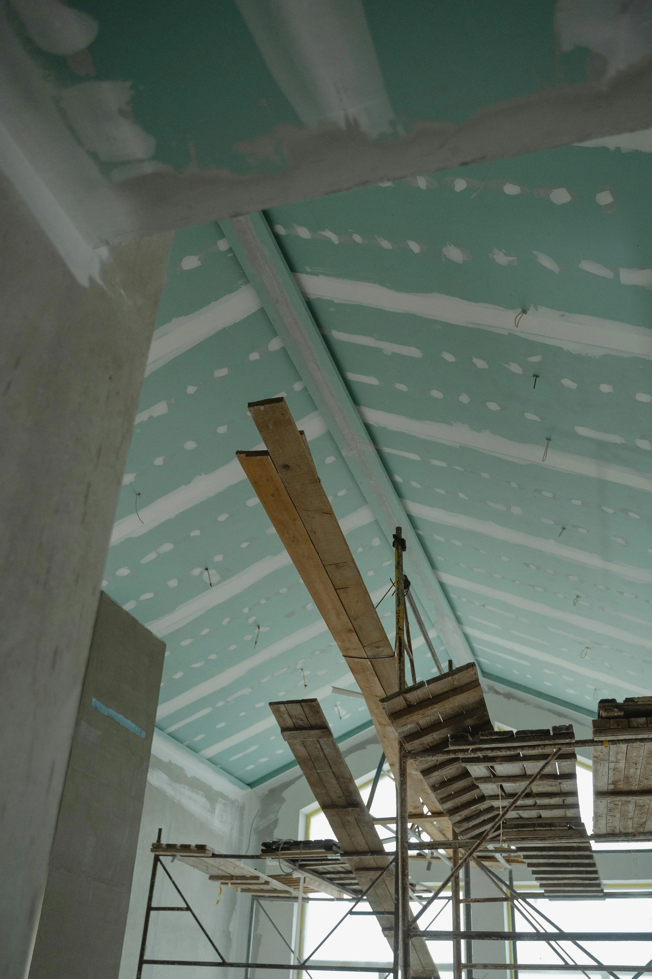 Interior of a building under construction with scaffolding, wooden planks, partial drywall or plasterboard, and a sloped ceiling with visible insulation or green drywall.