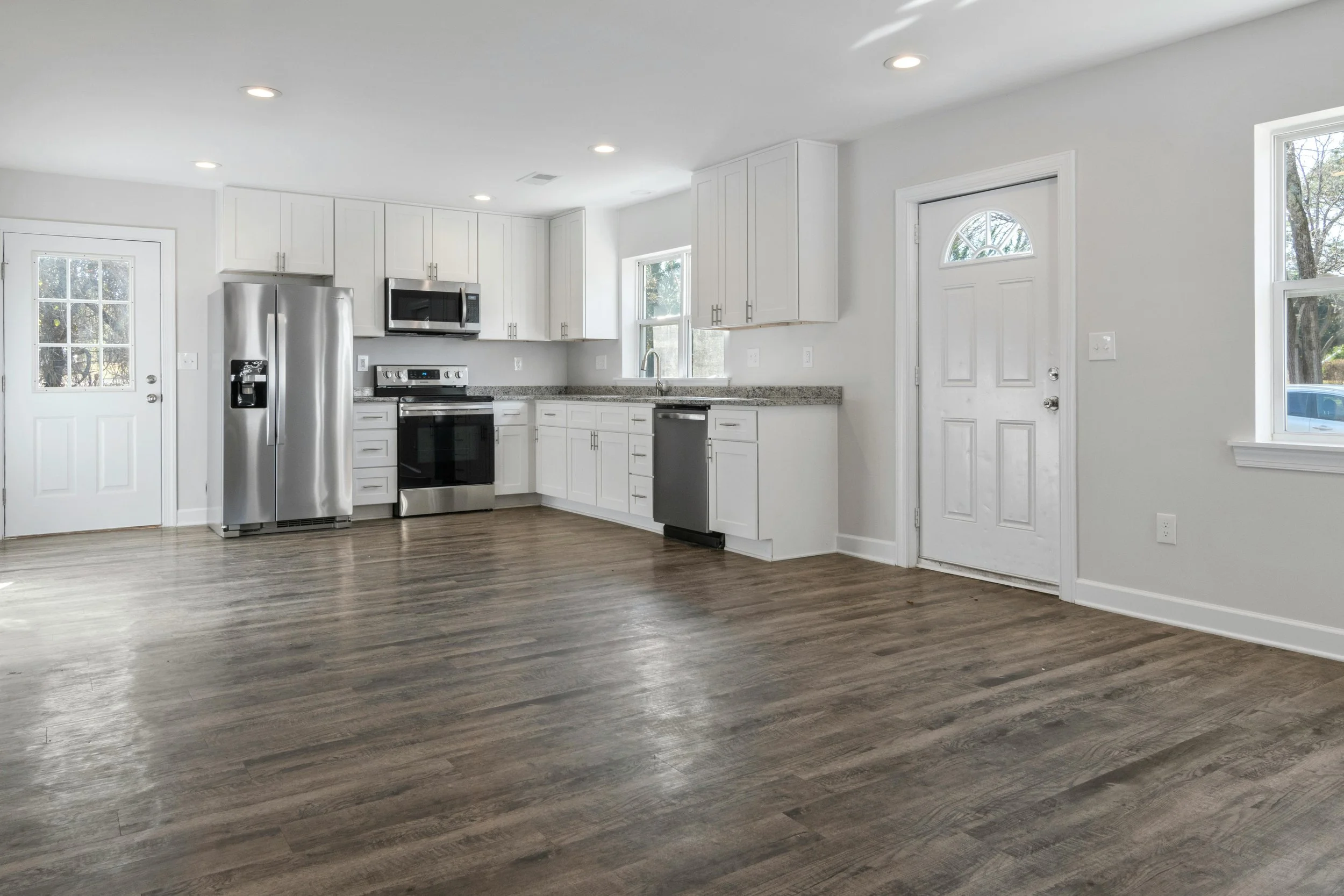 Empty kitchen with white cabinets, stainless steel refrigerator, microwave, oven, dishwasher, granite countertops, and hardwood floors, with windows and two exterior doors.