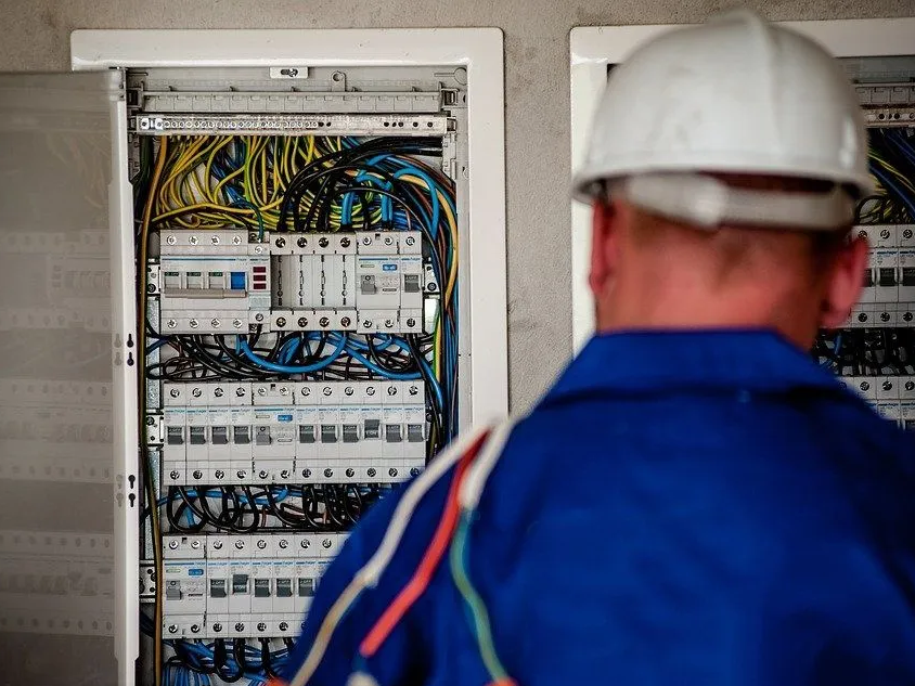 An electrician wearing a white hard hat and blue work clothes inspecting an open electrical panel with numerous wires and circuit breakers.