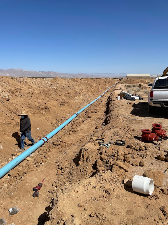 Construction workers installing a large blue pipeline in a dirt trench in a desert landscape.
