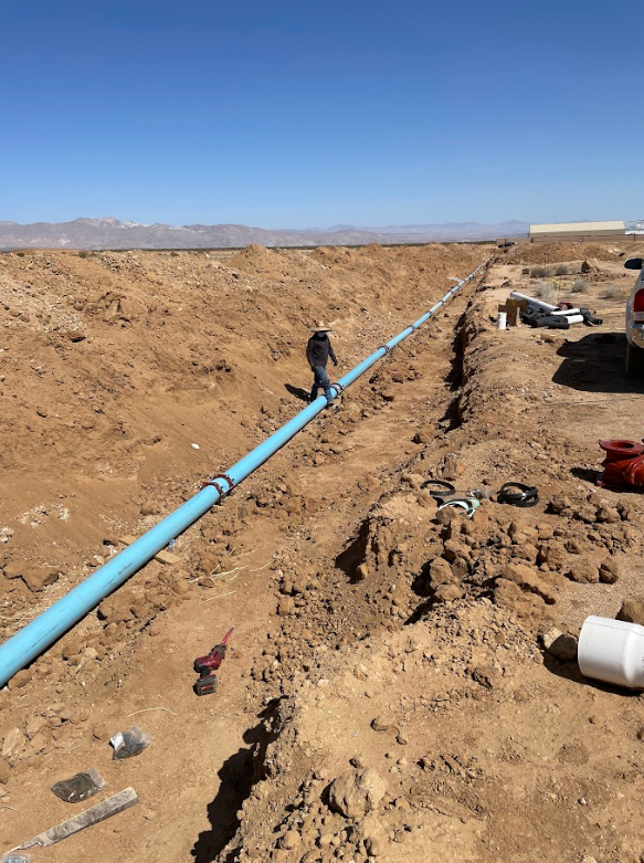 Construction worker walking along a large pipeline in a trench in an open desert landscape under a clear blue sky.