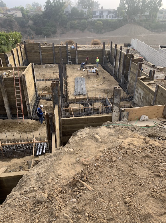 Construction site with workers assembling foundation walls using steel rebar, surrounded by dirt, with several houses and trees in the background.
