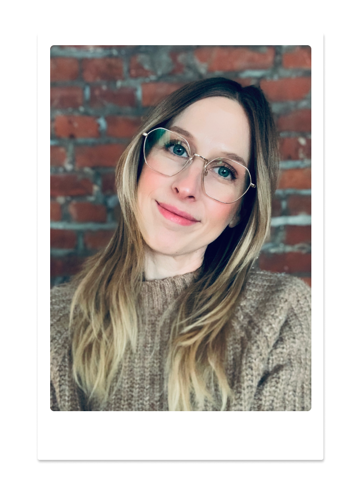 A woman with glasses and long wavy hair smiling in front of a red brick wall.