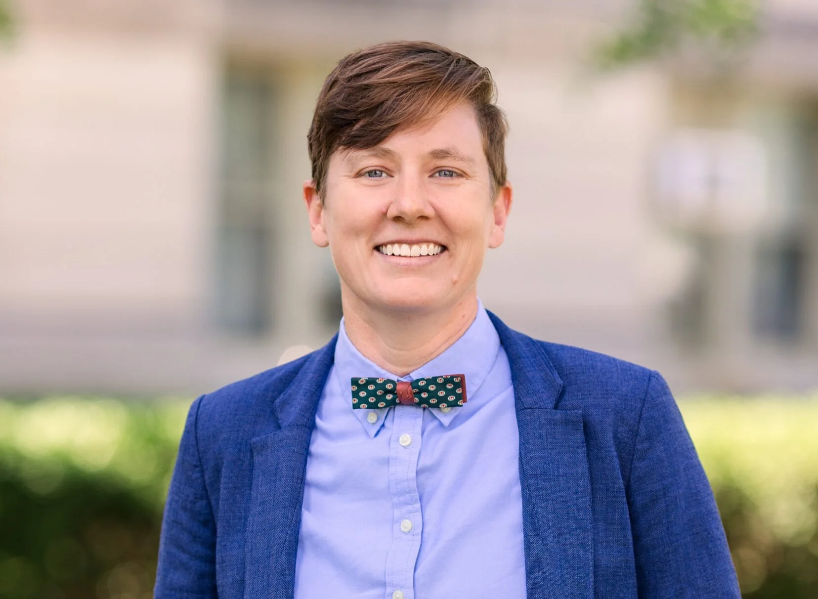 A smiling person with short brown hair, wearing a blue blazer, light blue shirt, and a green bow tie with a pattern, standing outdoors with a blurred background.