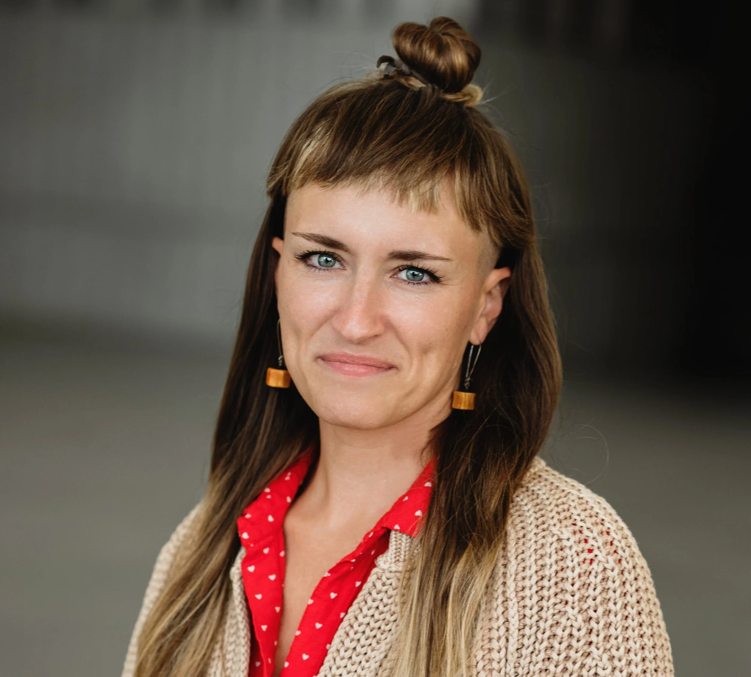 A woman with long brown hair and blue eyes, wearing a beige knit cardigan, red shirt with white hearts, and gold geometric earrings, standing in front of a neutral background.