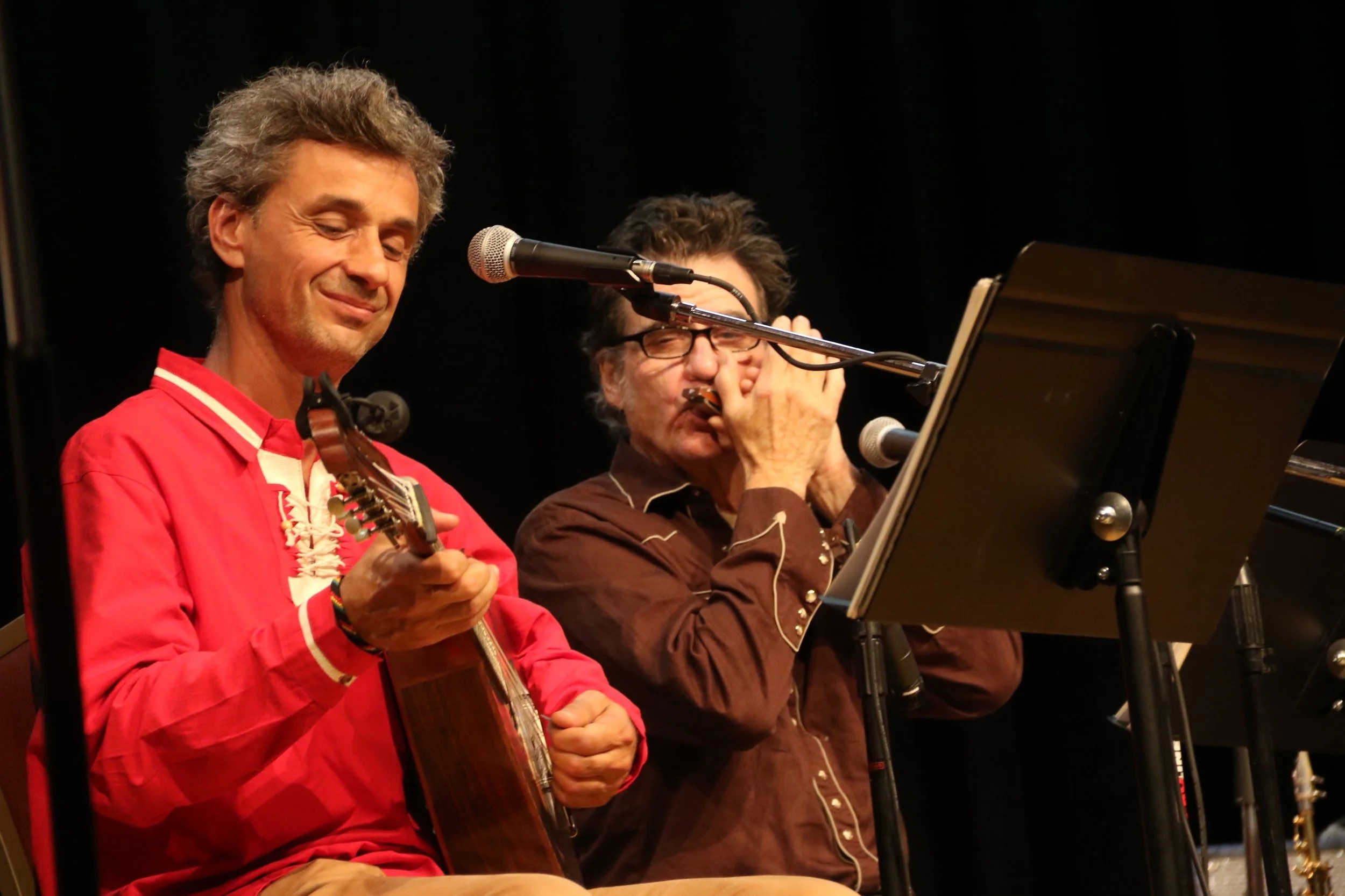 Two musicians performing on stage, one playing guitar and smiling, the other covering his ears while singing into a microphone.