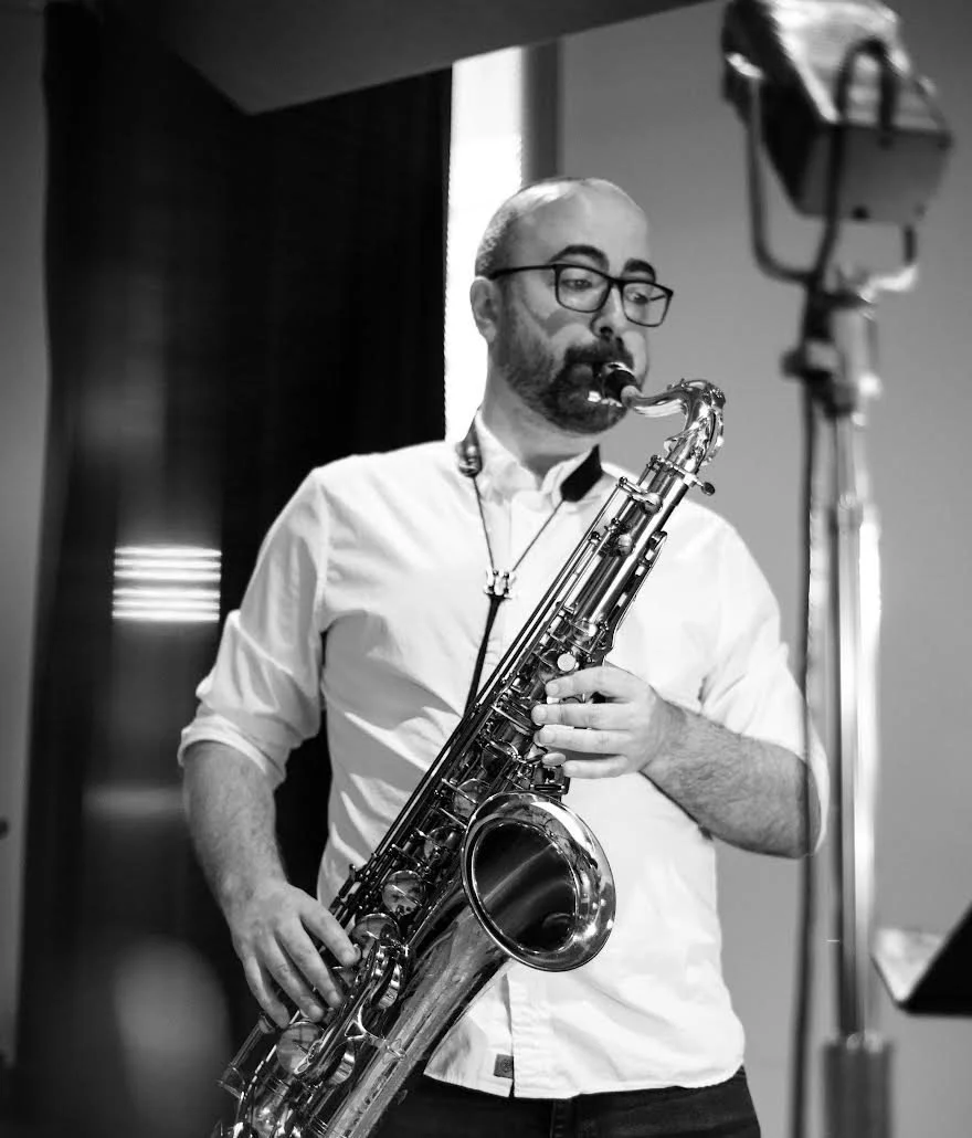 A man with glasses and a beard playing a saxophone in a recording studio.