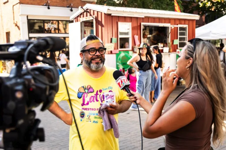 A man wearing glasses, a yellow Latino cultural festival T-shirt, and a hat is being interviewed by a woman holding a microphone. People are gathered in the background near a small colorful building at an outdoor event or street fair.