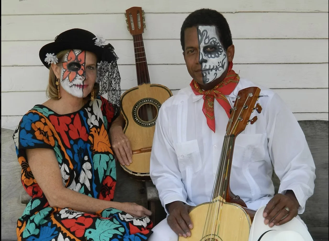 Two people dressed in colorful traditional Mexican attire with face paint, holding guitars, sitting against a white wooden wall.