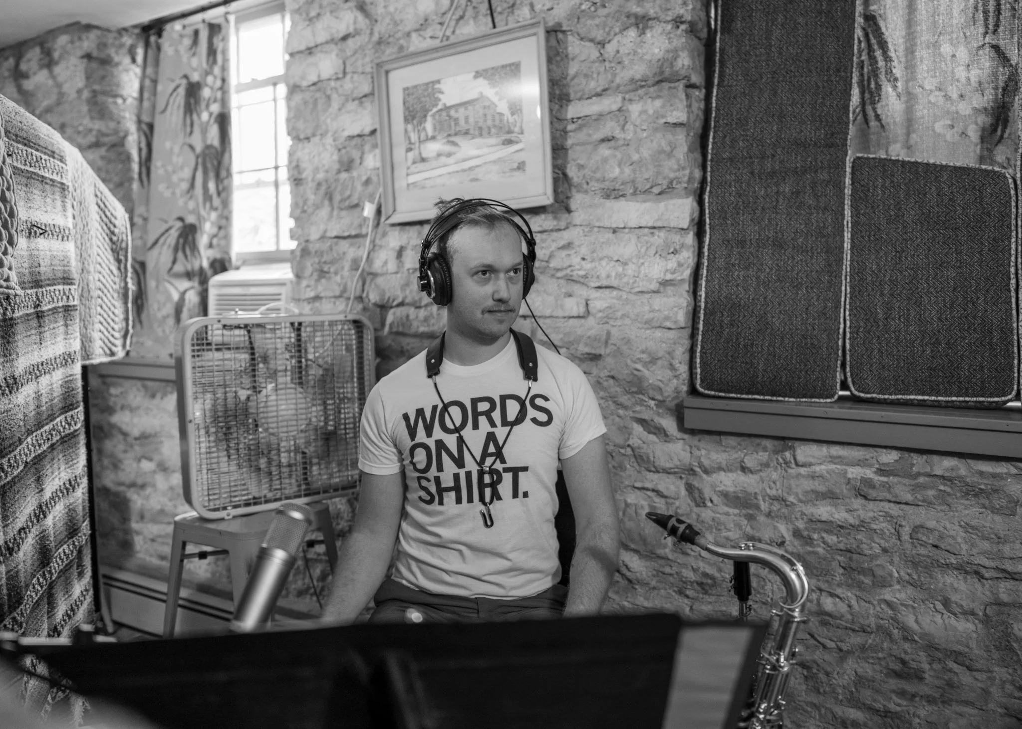 A saxophonist wearing headphones and a T-shirt that says 'Words on a Shirt' in front of a microphone and a music stand with a saxophone beside him.