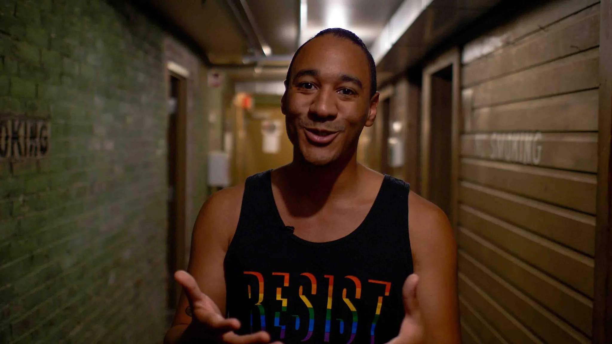 A person smiling and speaking in a hallway with brick and wooden walls, wearing a sleeveless shirt with a rainbow-colored 'RESIST' slogan.