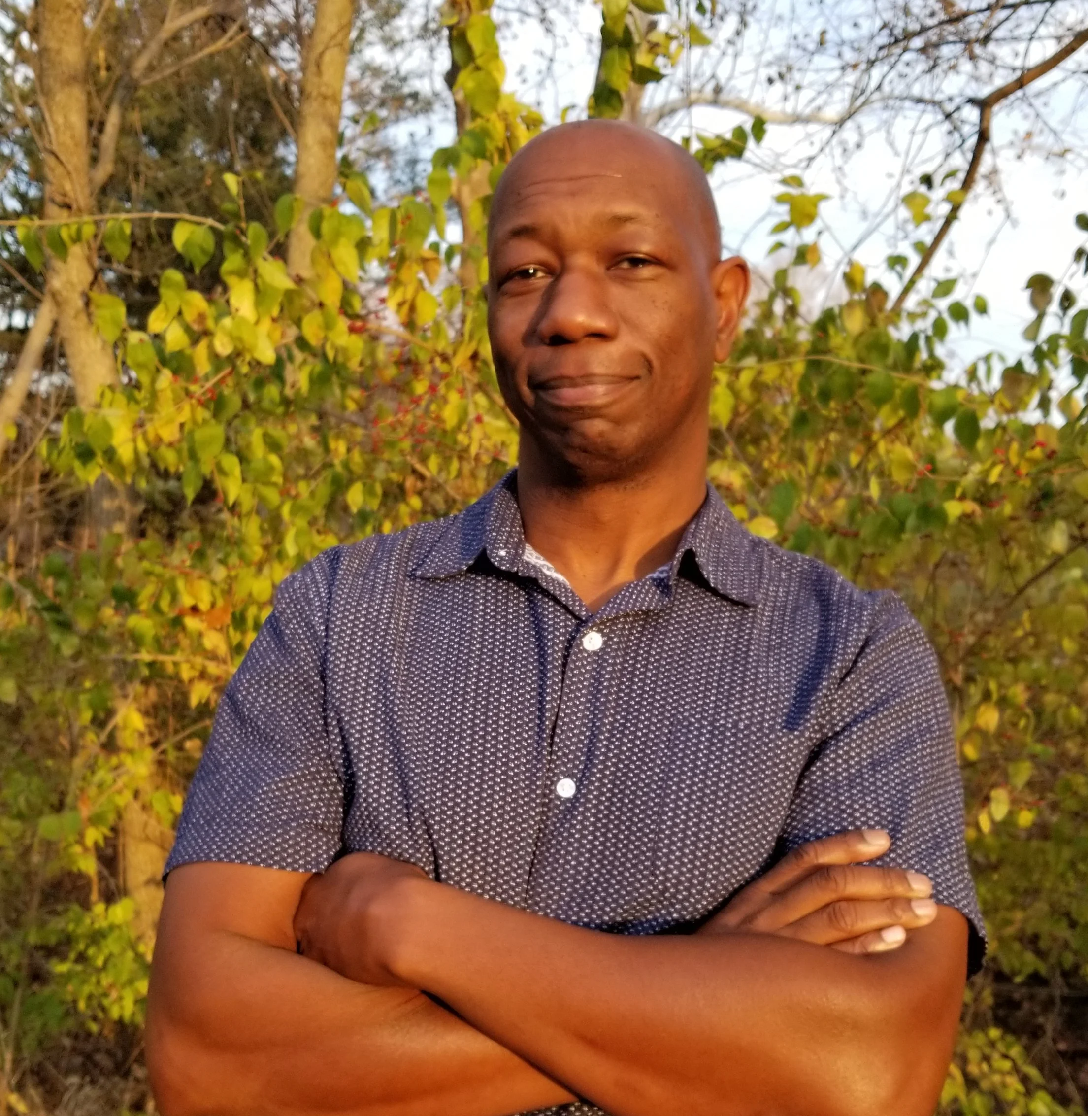 A man with crossed arms standing outdoors in front of trees with green and yellow leaves, in sunlight.