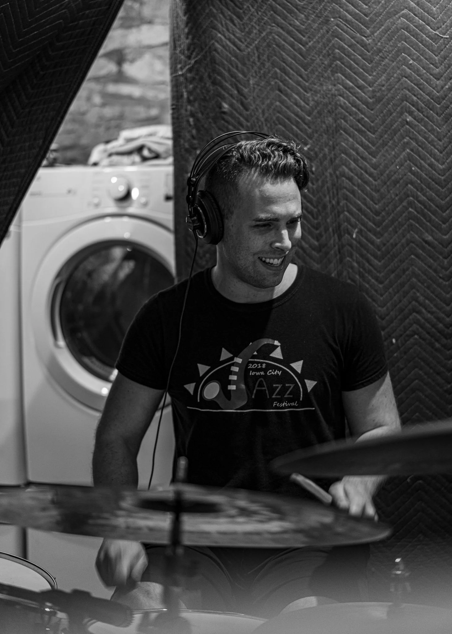 A drummer wearing headphones and a T-shirt with '2018 Iowa City Jazz Festival' is playing drums in a room with soundproofing panels, with a washing machine visible in the background.