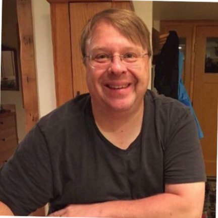 A smiling man with glasses and light brown hair, wearing a black T-shirt, sitting indoors with a wooden door and a coat hanging in the background.