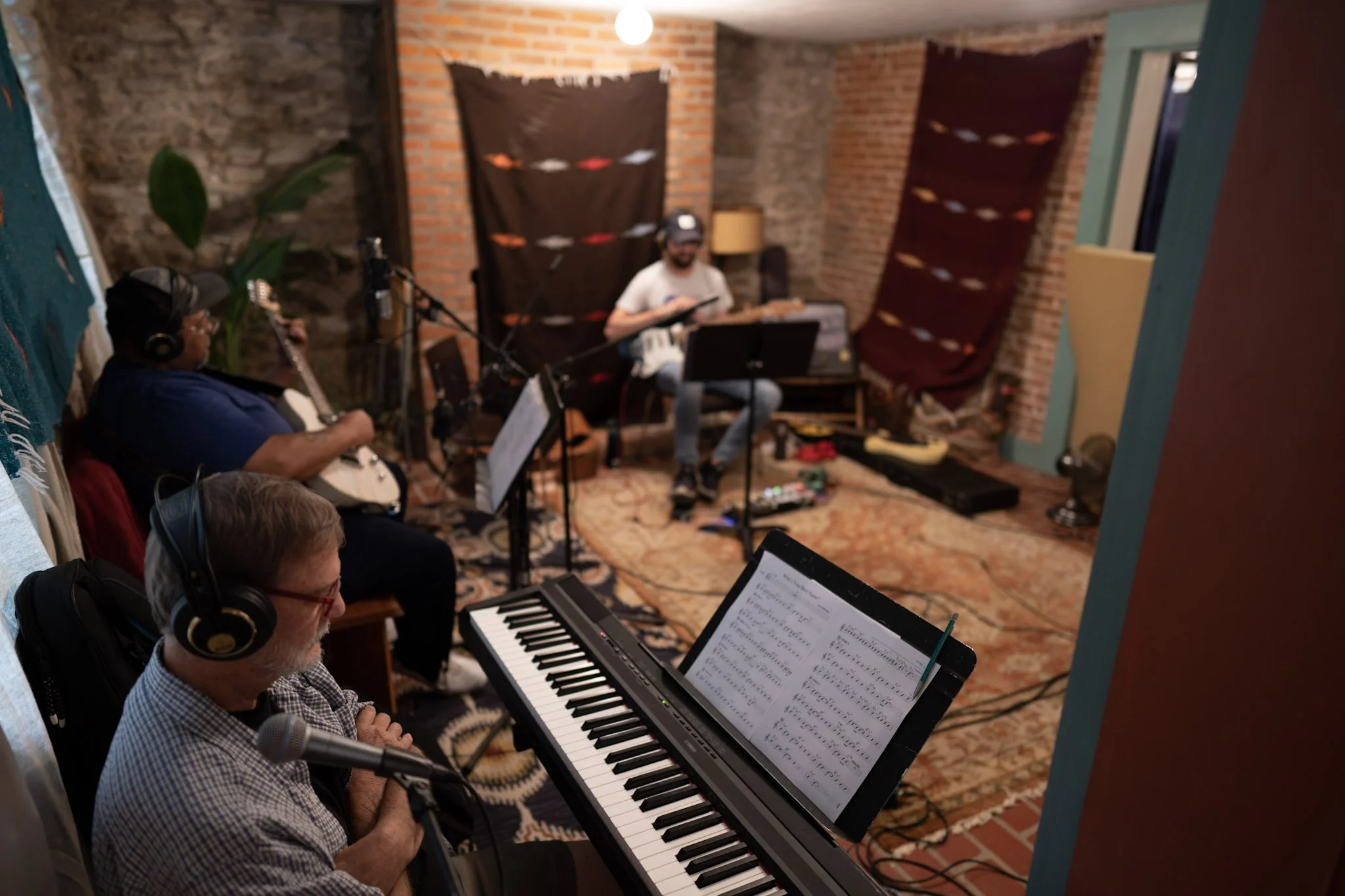 A group of musicians in a recording studio, with a man at a keyboard reading sheet music, another man playing an electric guitar, and a person in the background reading a tablet. The studio has brick walls, decorative fabric hanging on the walls, and equipment on the floor.