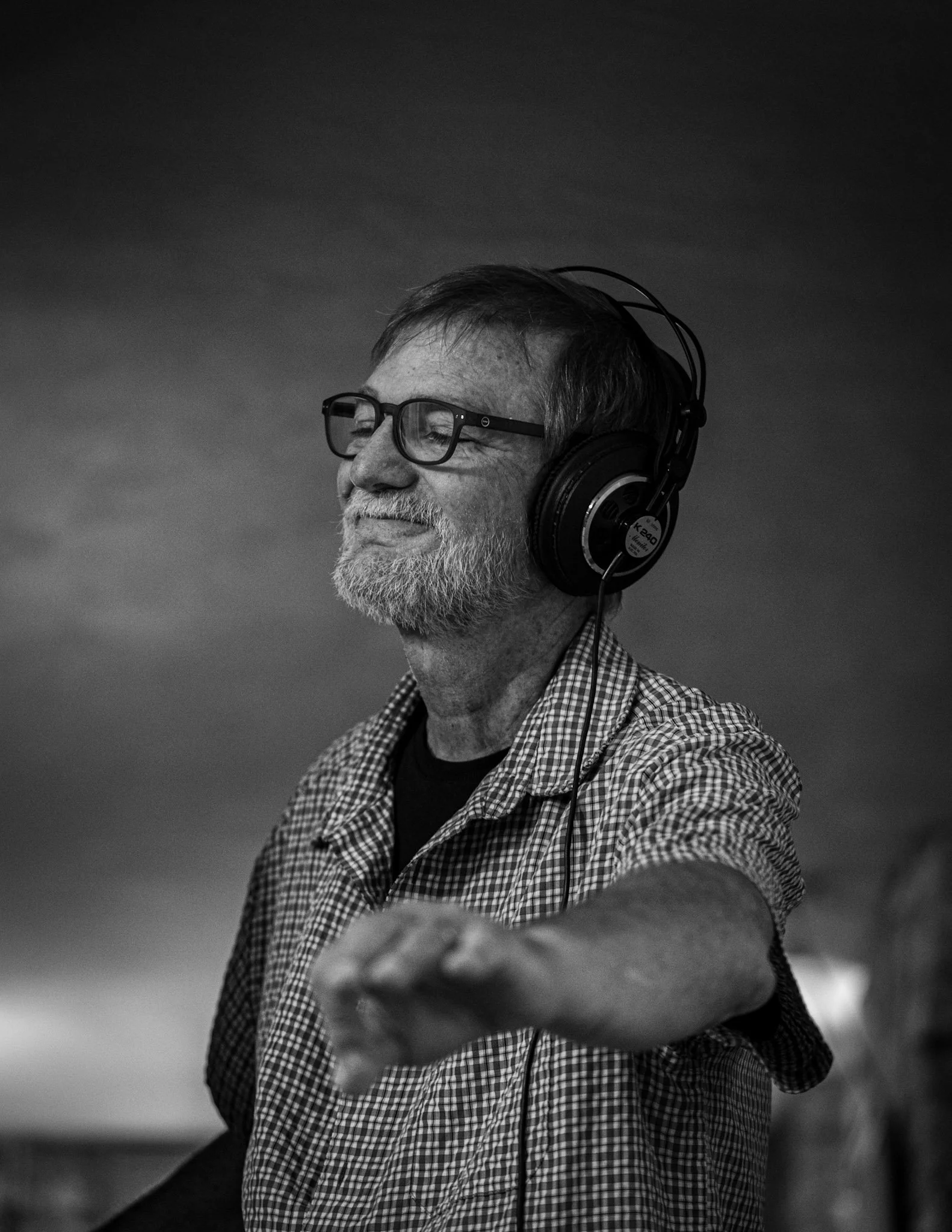 Older man with glasses, headphones, and a checkered shirt, smiling with eyes closed and reaching out