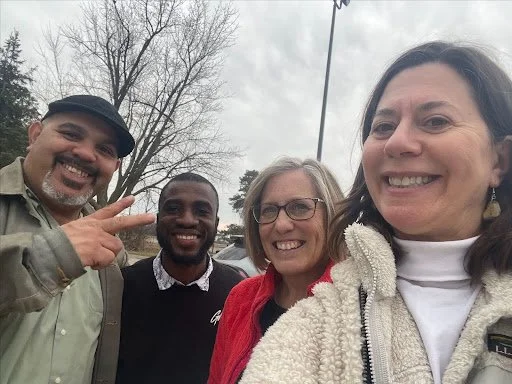 Four smiling people, including two men and two women, taking a selfie outdoors with trees and overcast sky in the background.