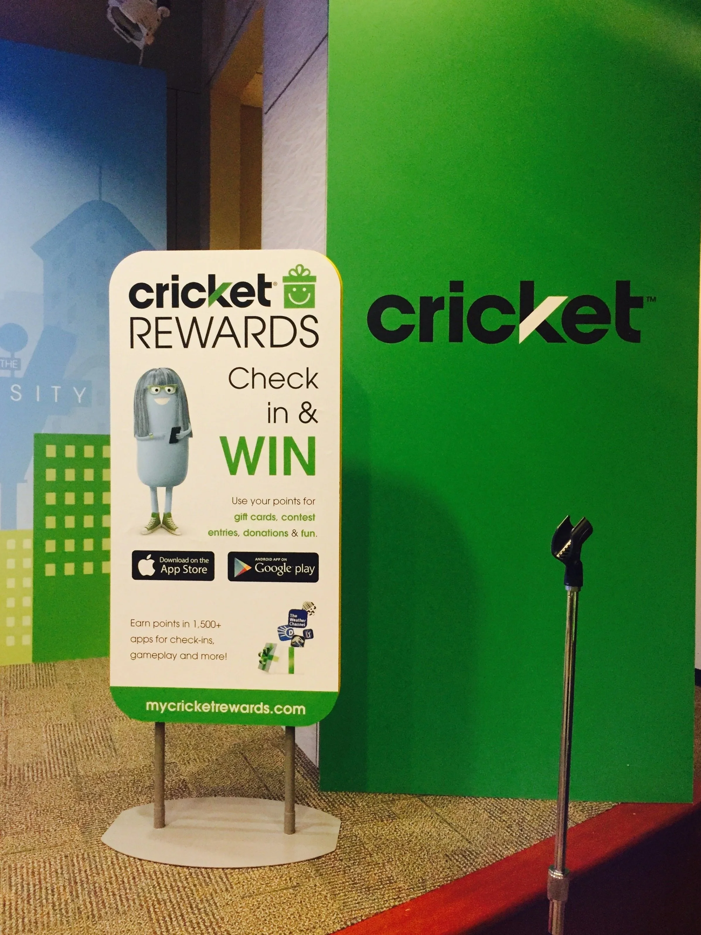 Sign promoting Cricket Rewards with a cartoon character mascot, directions to check in and win prizes through an app available on the App Store and Google Play, and a mic stand next to a green Cricket backdrop.