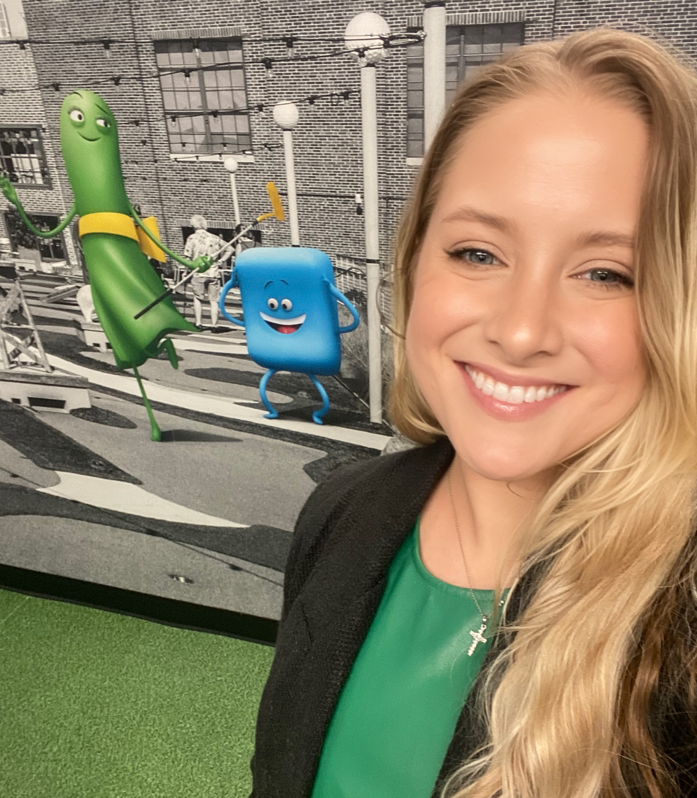 A smiling woman with blonde hair taking a photo in front of a colorful animated billboard showing cartoon objects on a city street.