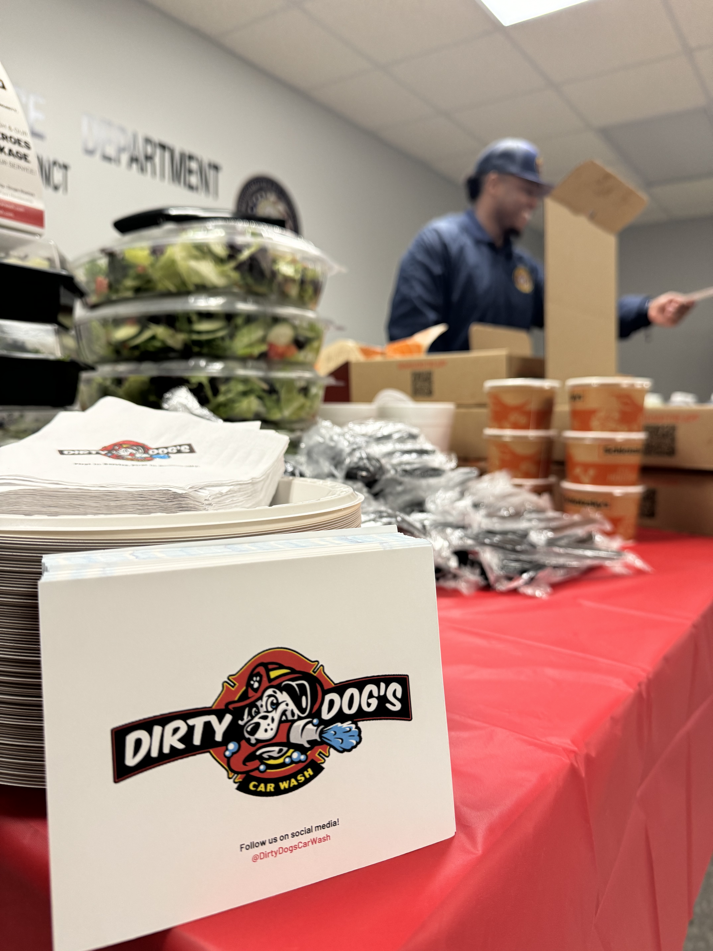A table with food and supplies at 'Dirty Dog's Car Wash' event, showing a sign with the logo, stacks of salad, and cups, with a person in uniform in the background.