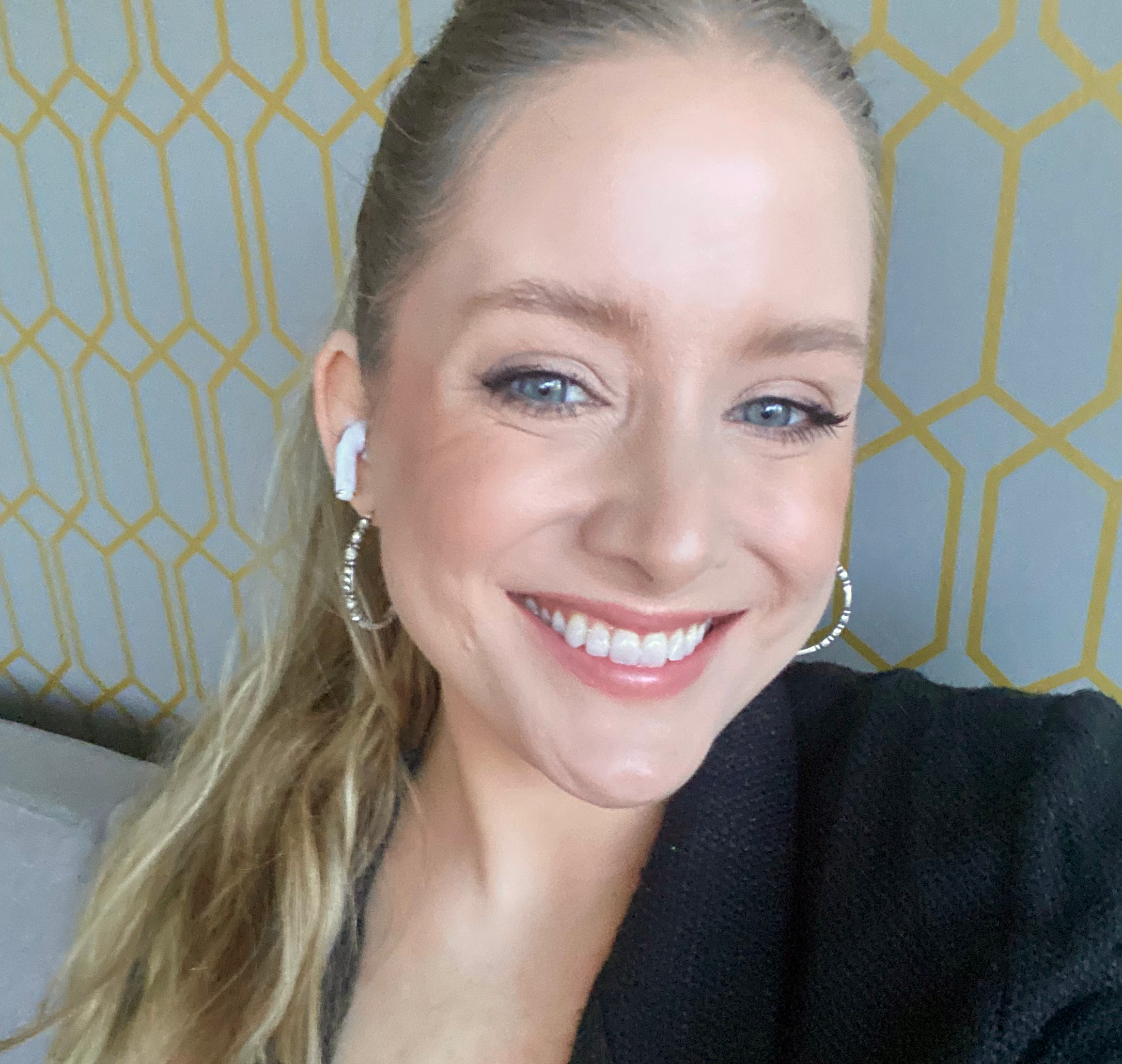 A woman with blonde hair and blue eyes smiling, wearing hoop earrings and a black top, with a yellow patterned background.