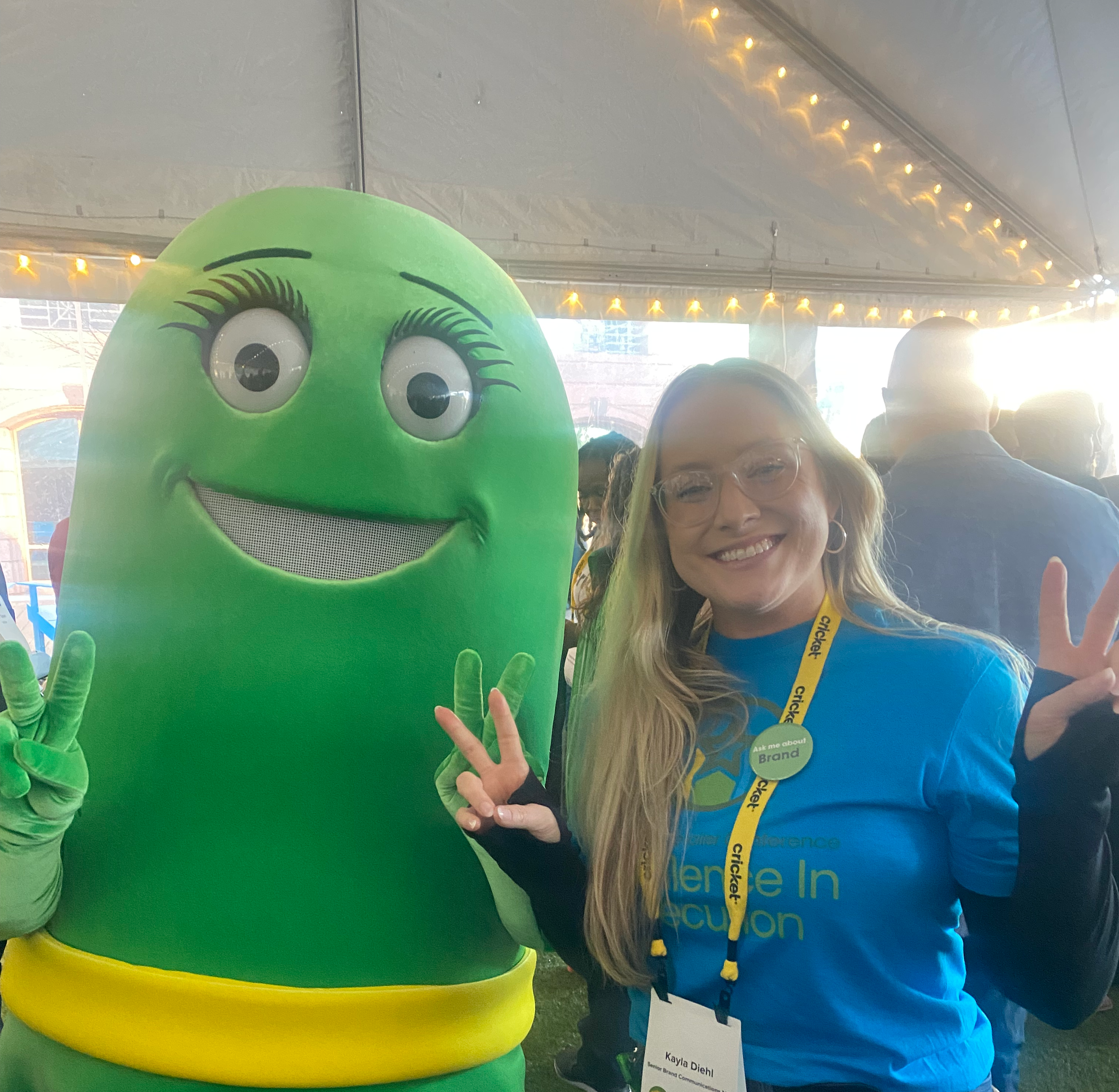 A woman smiling and wearing glasses, a blue T-shirt, and an event badge and lanyard, standing next to a large green mascot with big eyes, a wide smile, and two fingers raised in a peace sign inside a tent with lights overhead.