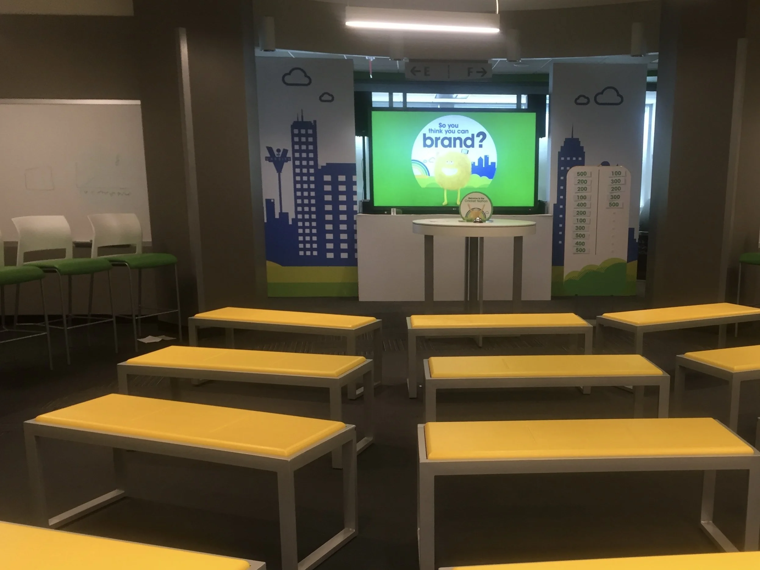 Empty room with yellow benches, white chairs with green seats, and a large screen displaying a colorful presentation in a corporate or conference setting.