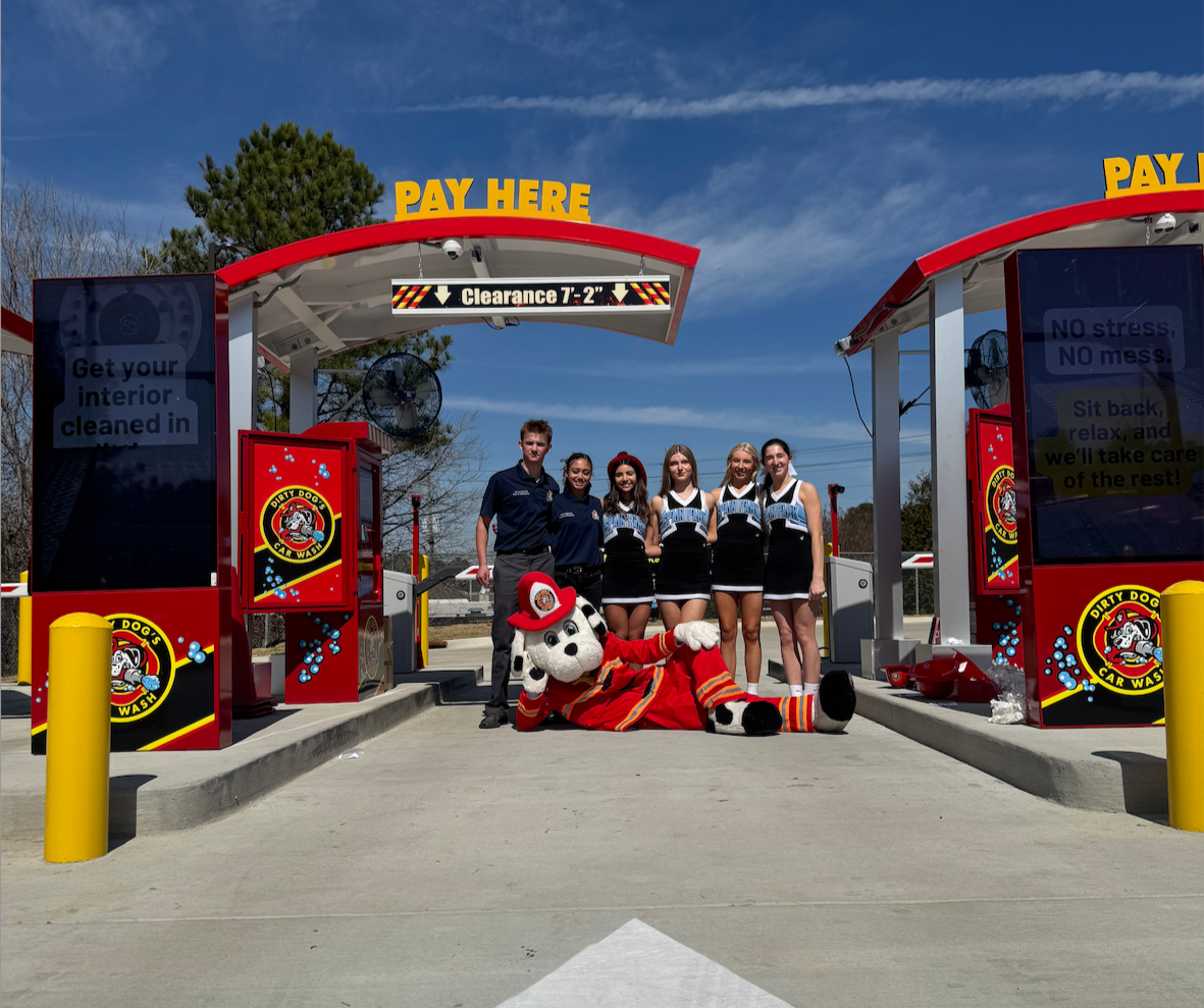Group of people and a mascot at a car wash with bright red wash stations, a large sign that reads 'Pay Here,' and clear blue sky.