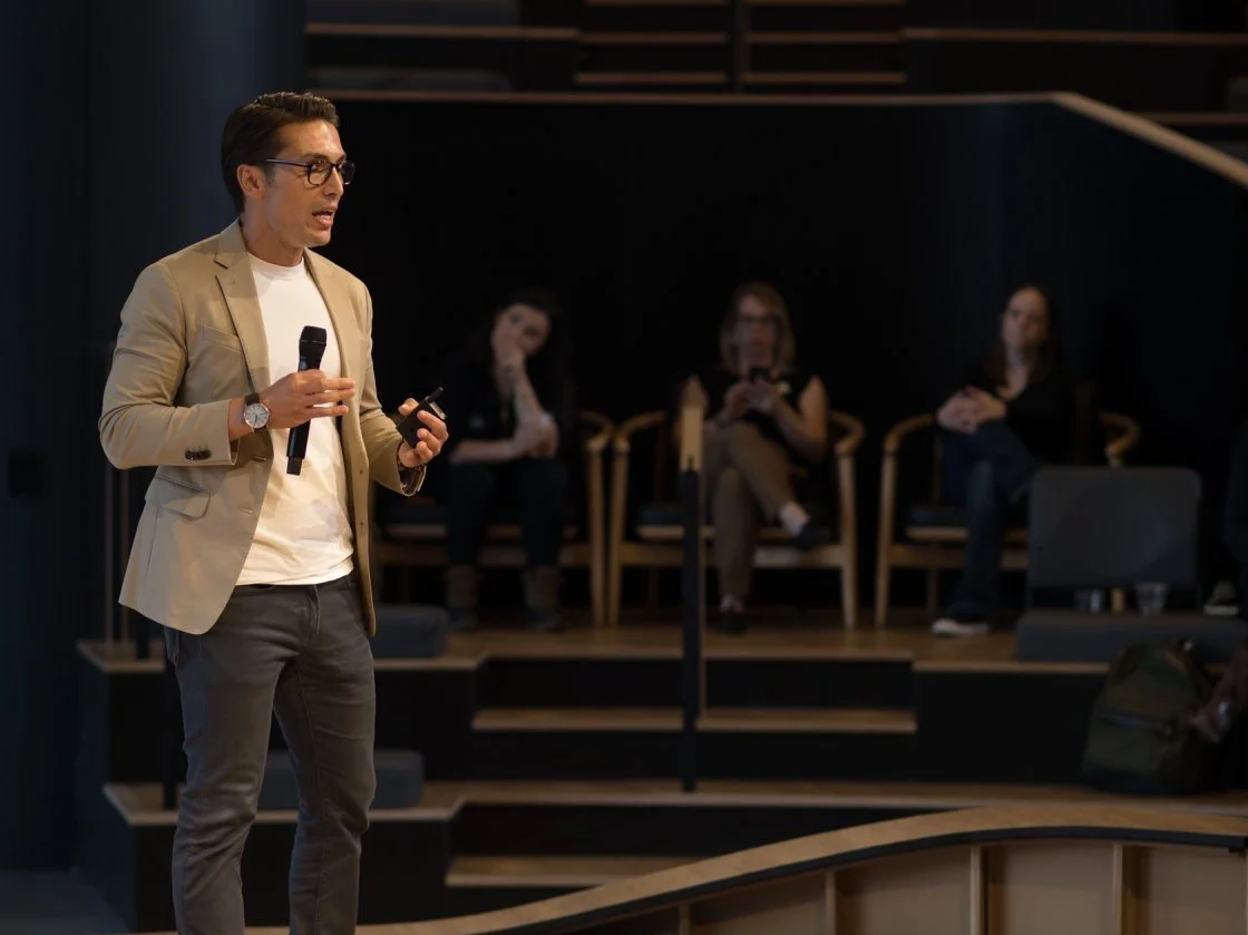 A man in a tan blazer and glasses giving a presentation with a microphone in hand, standing on a stage with four seated women in the background.