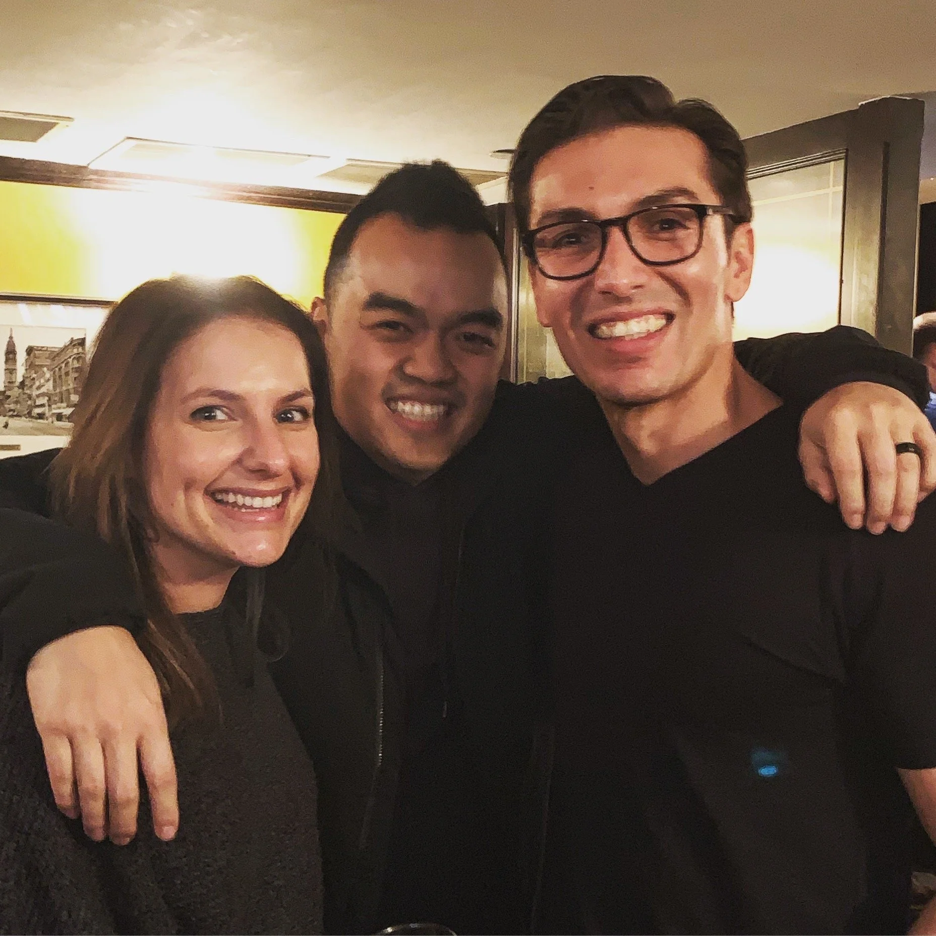 Three people standing close together with their arms around each other, smiling at the camera in a warmly lit indoor setting.