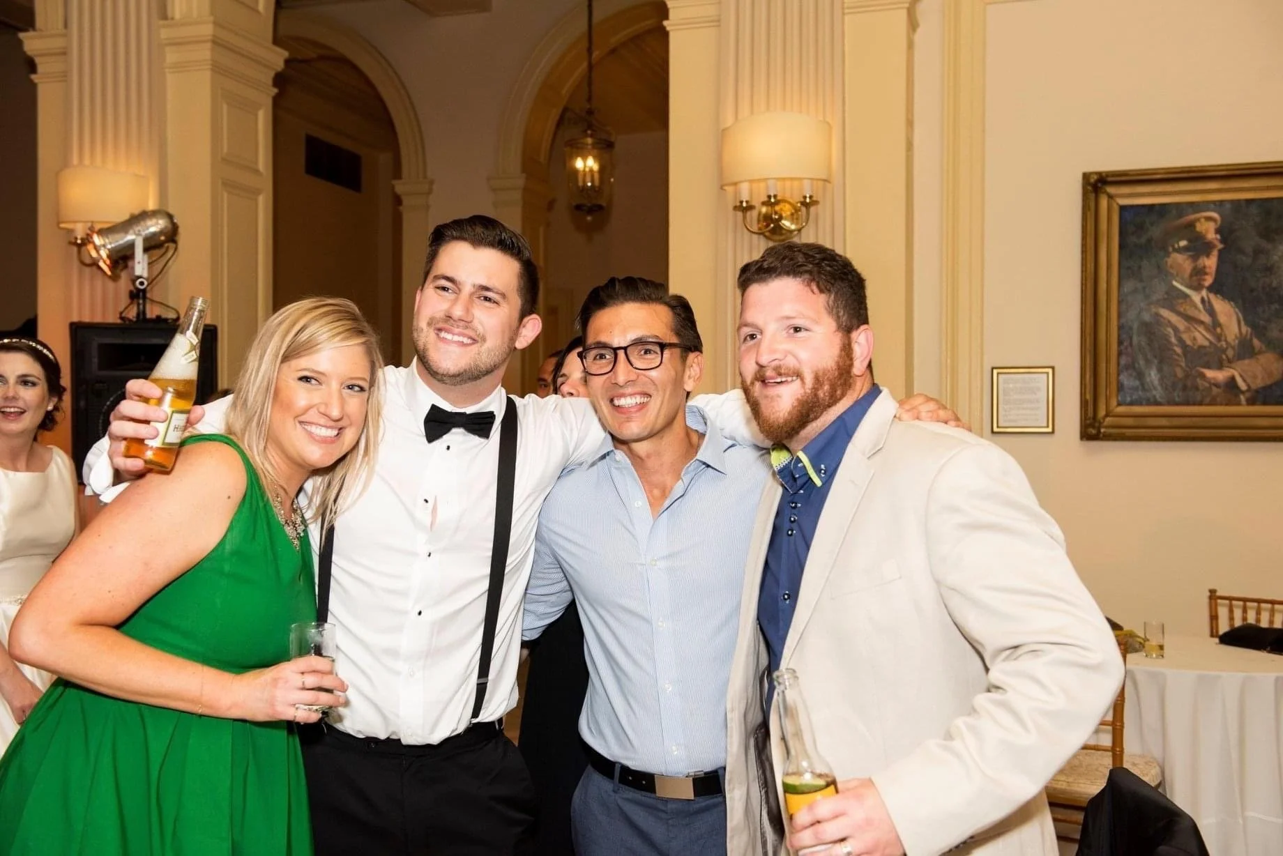 Group of five people at a party, smiling and holding drinks, dressed in formal attire, standing in a decorated room with framed pictures and lamps.