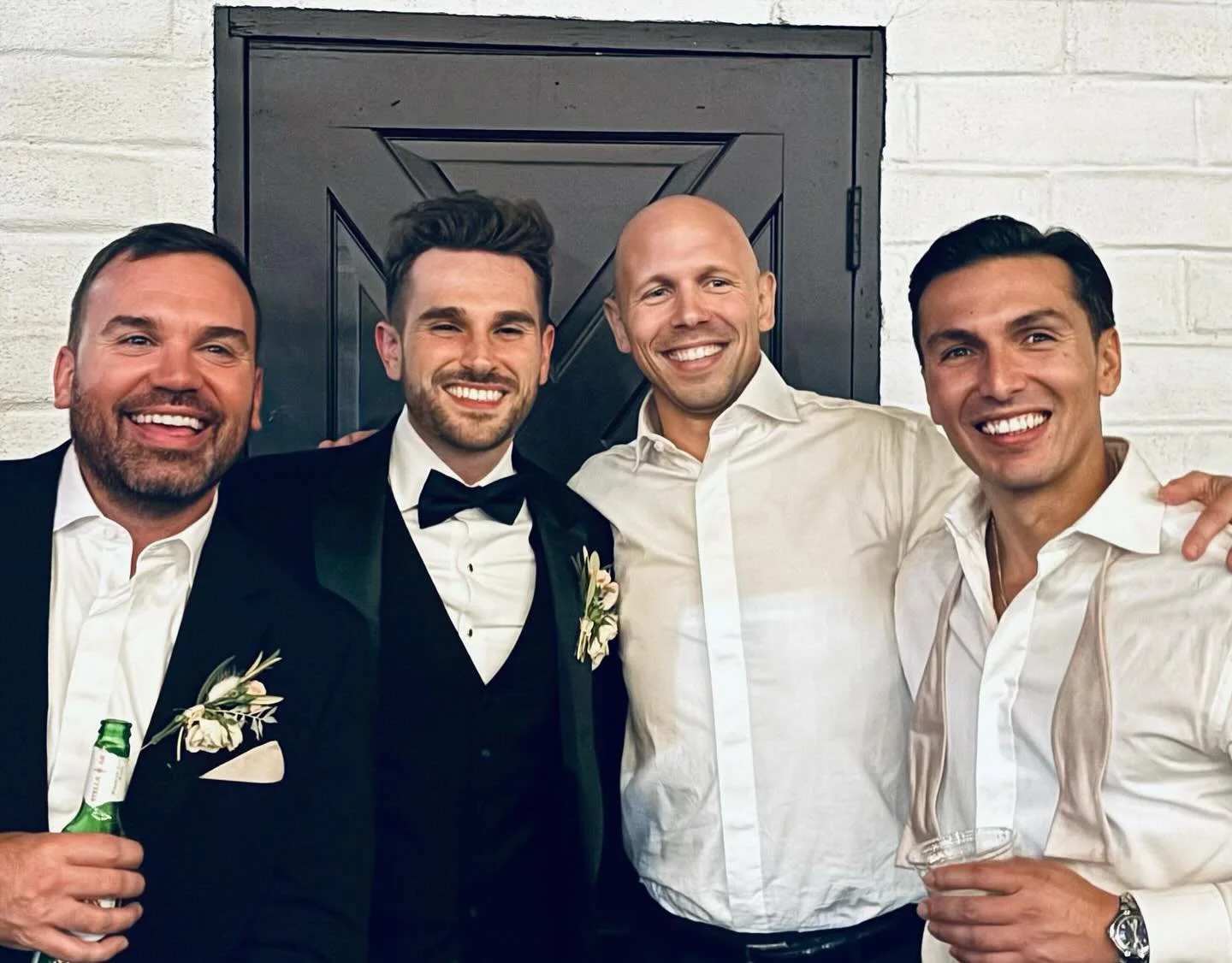 Four men smiling at a celebration event, everyone dressed in formal attire, with one holding a beer bottle and another holding a glass, standing in front of a dark wooden door and white brick wall.
