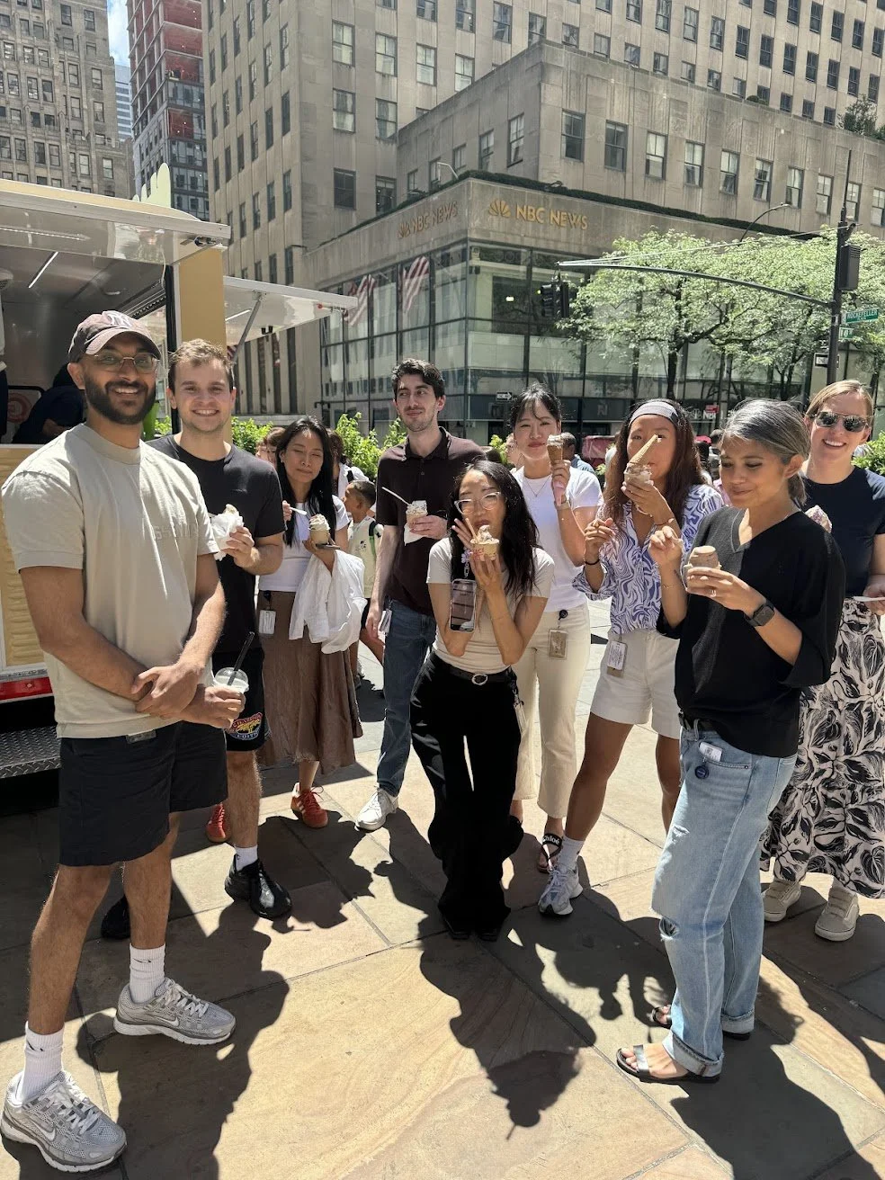 A group of people enjoying ice cream outdoors on a city sidewalk with tall buildings and trees in the background.